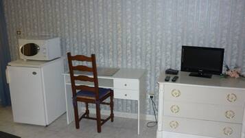 Une pièce de taille modeste, avec un bureau blanc, une chaise en bois, un petit meuble blanc avec des tiroirs, un téléviseur et un four à micro-ondes posé sur un petit appareil blanc.