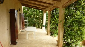 A covered outdoor area with a white table and chairs, surrounded by stone flooring and greenery. - Domaine de la Rode Buze (Cahors)