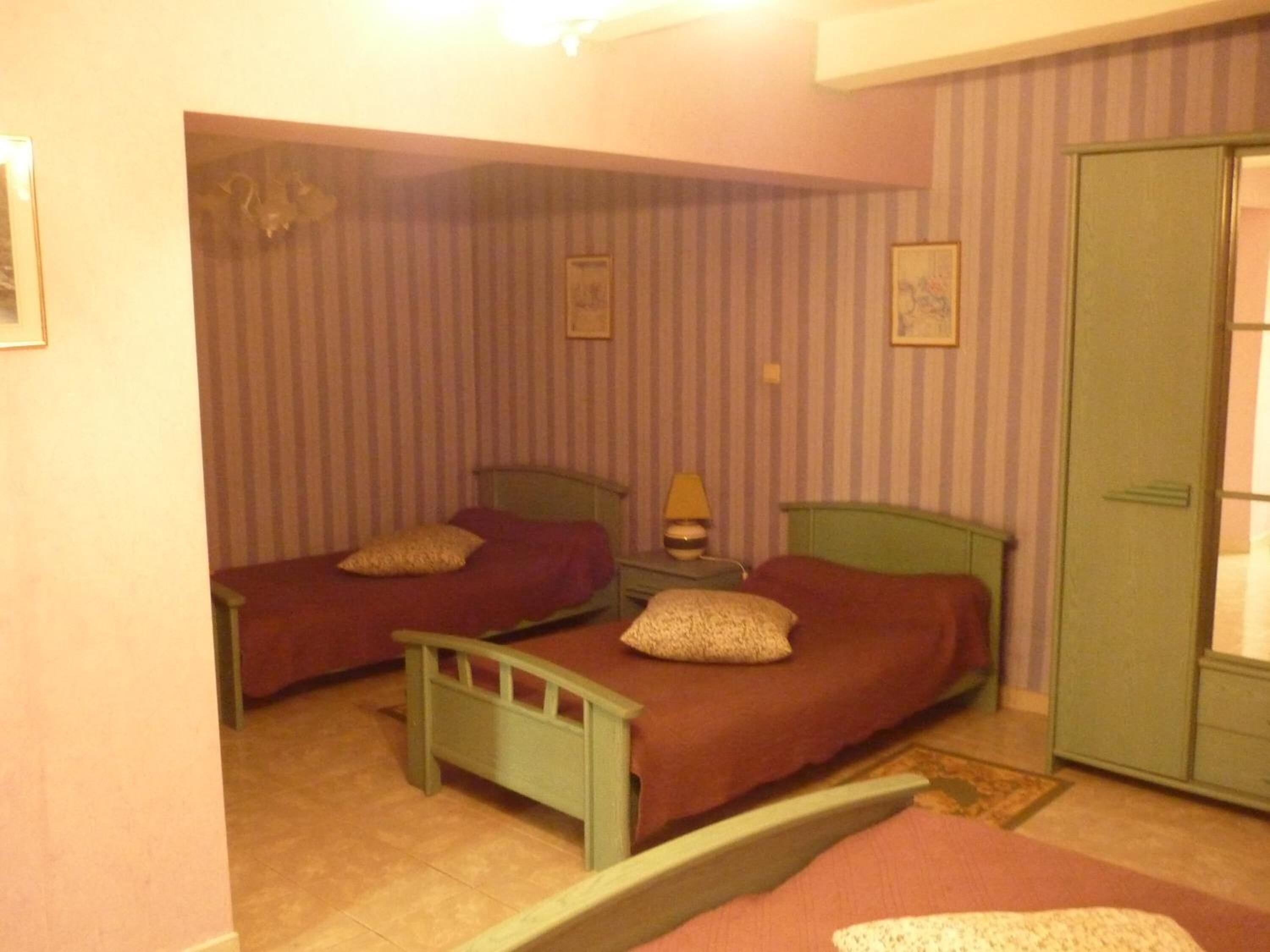 A room with two beds, striped wallpaper, a green wardrobe, and a small bedside table with a lamp.