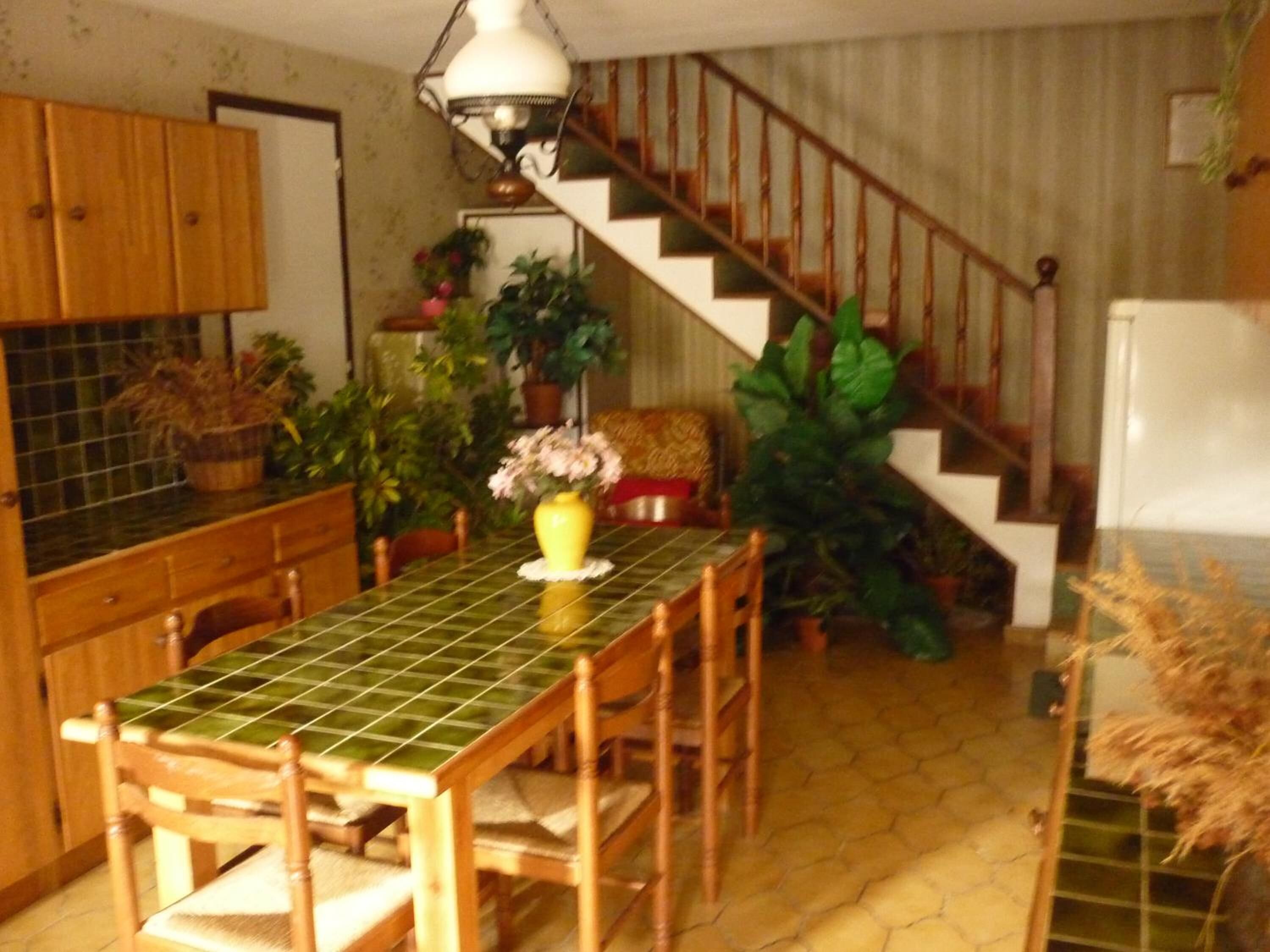 A kitchen with a tiled table, wooden chairs, a staircase, and various potted plants.