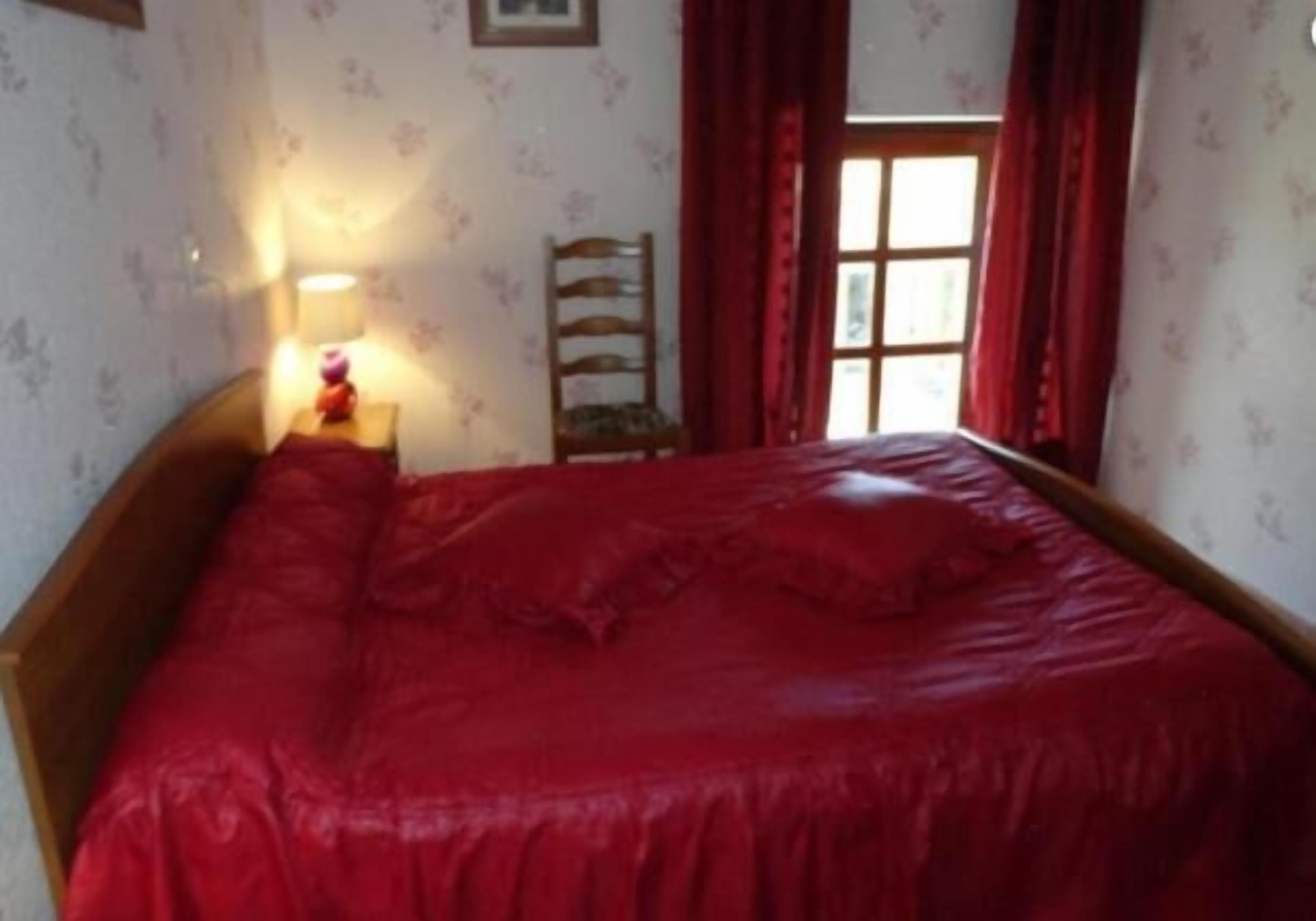 Een slaapkamer met een houten bed, een nachtkastje met een lamp en een raam met rode gordijnen.