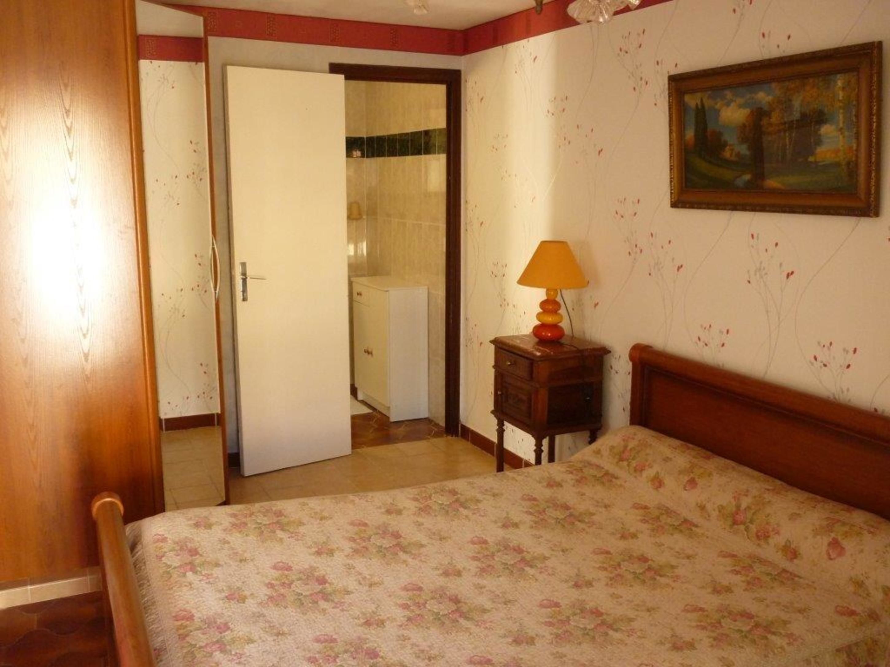 Un dormitorio con una cama de madera, una mesita de noche con una lámpara, una puerta que da a un baño y un cuadro en la pared.