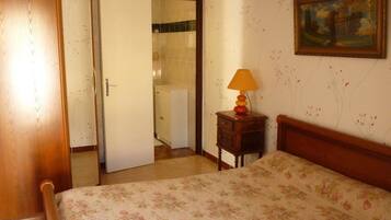 Un dormitorio con una cama de madera, una mesita de noche con una lámpara, una puerta que da a un baño y un cuadro en la pared.