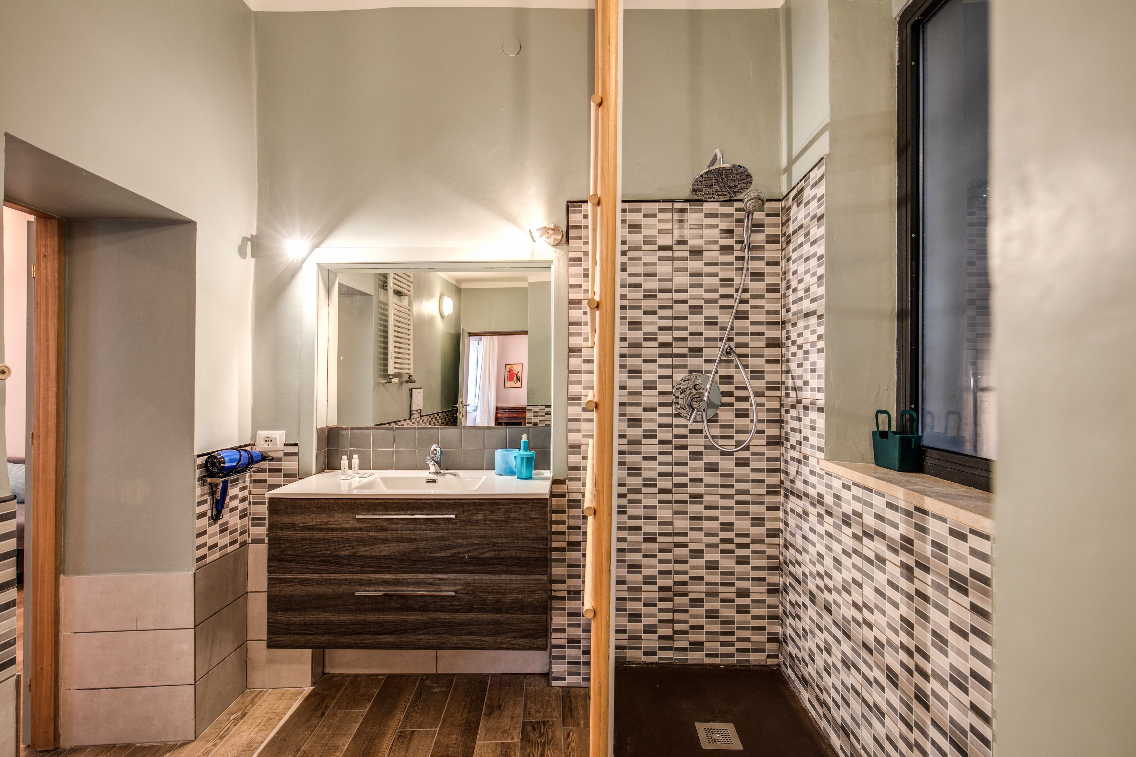 Une salle de bain moderne avec un grand miroir, un meuble-lavabo en bois, une douche avec un mur carrelé à motifs et une fenêtre.