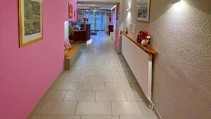 Een gang met roze wanden, een betegelde vloer en ingelijste foto's. Er is een balie met een spiegel en een plank met decoratieve objecten.