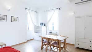 Ein Zimmer mit Esstisch, Stühlen, einem weißen Kleiderschrank und zwei mit Vorhängen versehenen Fenstern.