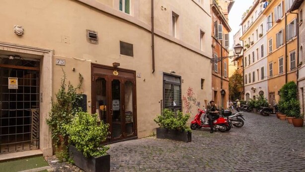 A narrow cobblestone street with buildings on either side, a closed metal gate, and a red scooter parked on the right. - Cappellari 4 Campo de Fiori (Rome)