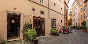 Une ruelle étroite pavée de pierres, bordée de bâtiments des deux côtés, avec un portail métallique fermé et un scooter rouge garé sur le côté droit.