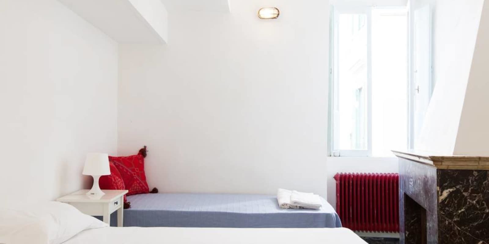 Quarto com cama, mesa de cabeceira, travesseiro vermelho, abajur branco e radiador.
