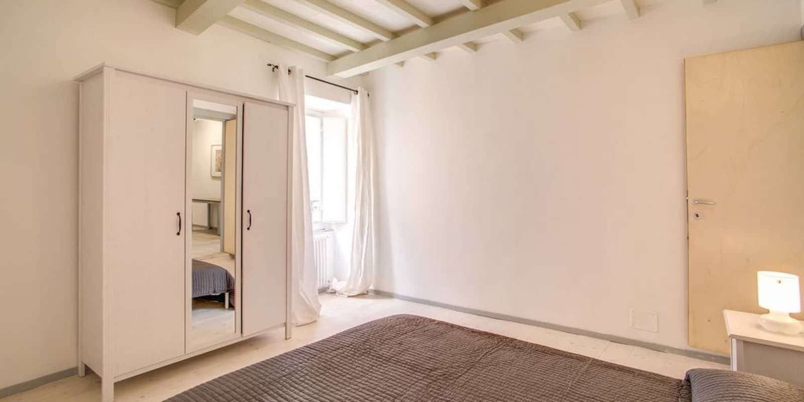 Un dormitorio con un armario grande, una cama con una manta marrón, una mesita de noche con una lámpara y una ventana con cortinas.