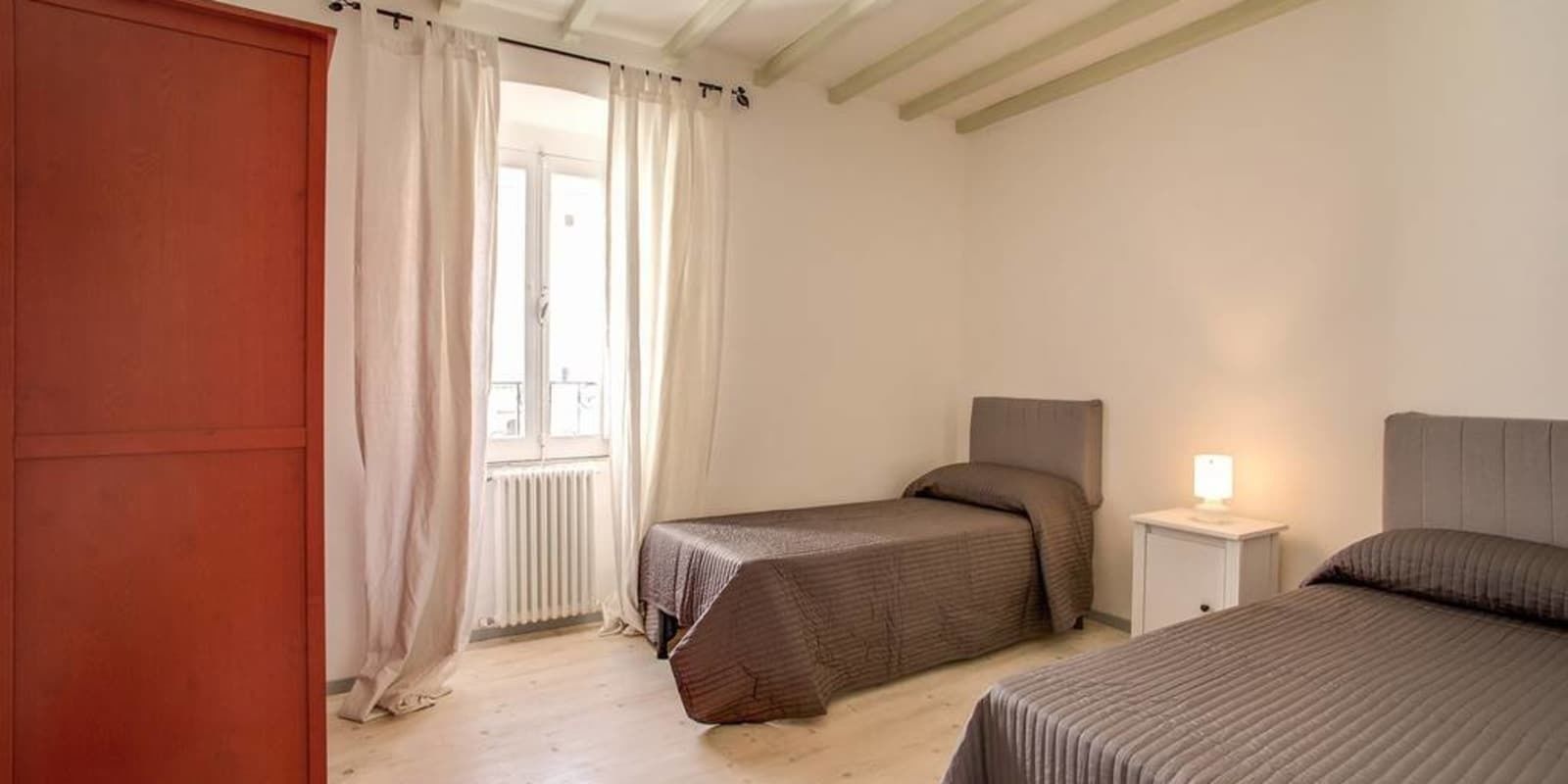 Ein Schlafzimmer mit zwei Einzelbetten, einem Nachttisch mit Lampe, einem Fenster mit Vorhängen und einem Heizkörper.
