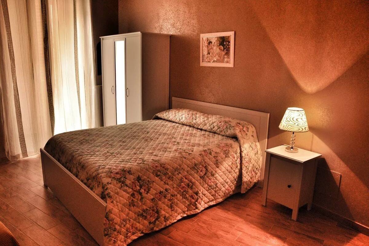 A bedroom with a bed, a nightstand with a lamp, a wardrobe, and a framed picture on the wall.