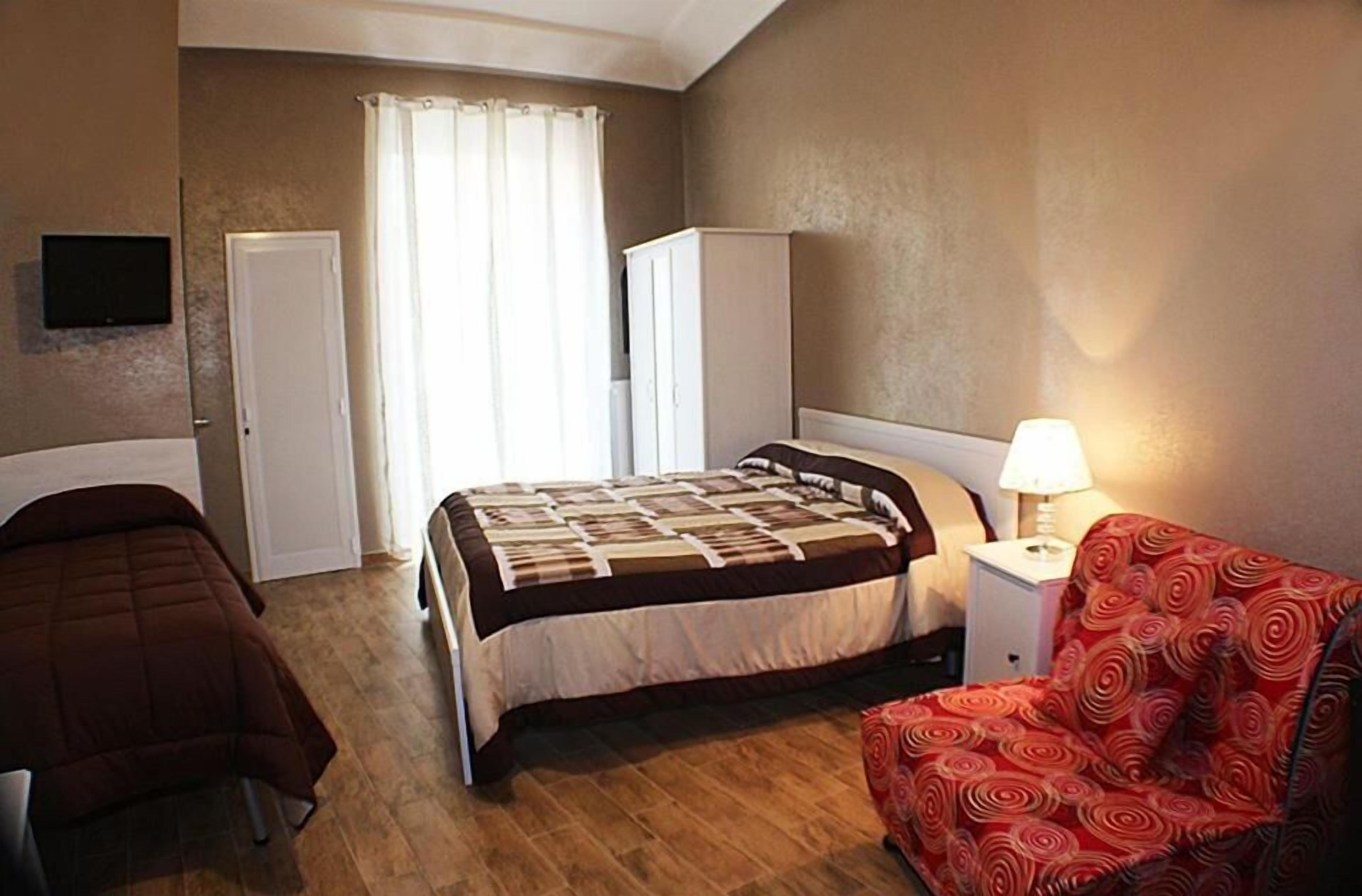 A hotel room with a bed, a chair, a nightstand with a lamp, a wardrobe, and a television on the wall.