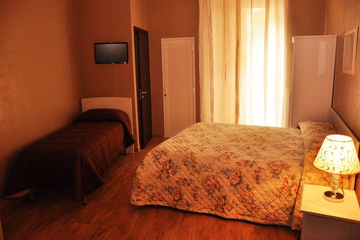 A bedroom with a bed, a nightstand with a lamp, a television on the wall, and a door.