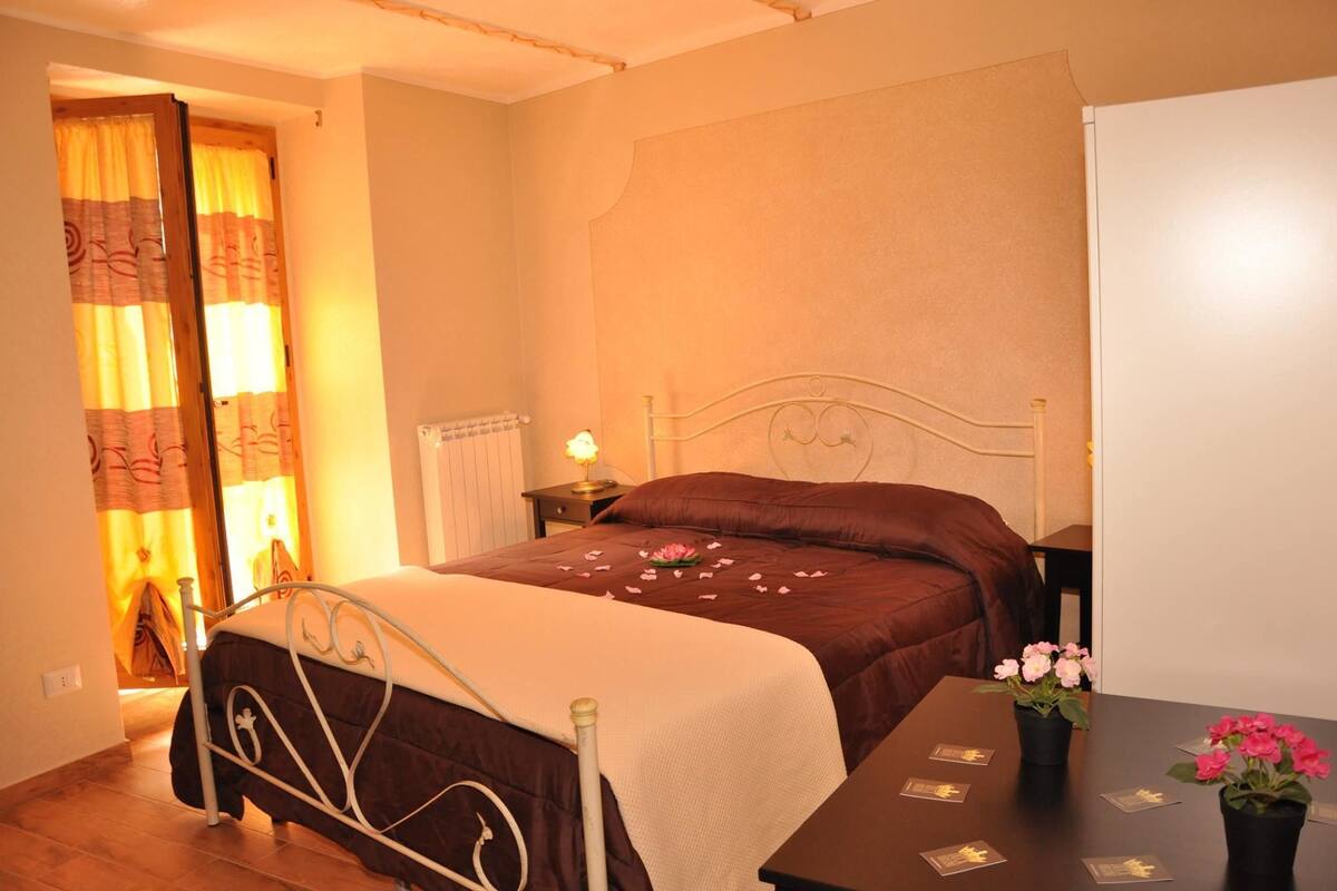 A bedroom with a bed, a nightstand, a table with a vase of flowers, and a door with curtains.