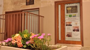 A hotel entrance with a wooden gate, a flower bed with pink and yellow flowers, and a signboard with hotel information. - B&B Duca Orsini (Gravina in Puglia)