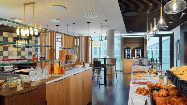 Un buffet de petit-déjeuner proposant une variété d’aliments et de boissons, un espace détente avec des chaises, et une vue sur la ville grâce à de grandes fenêtres.