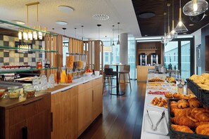 Buffet de café da manhã com diversos alimentos e bebidas, área de estar com cadeiras e vista para a cidade através de grandes janelas.
