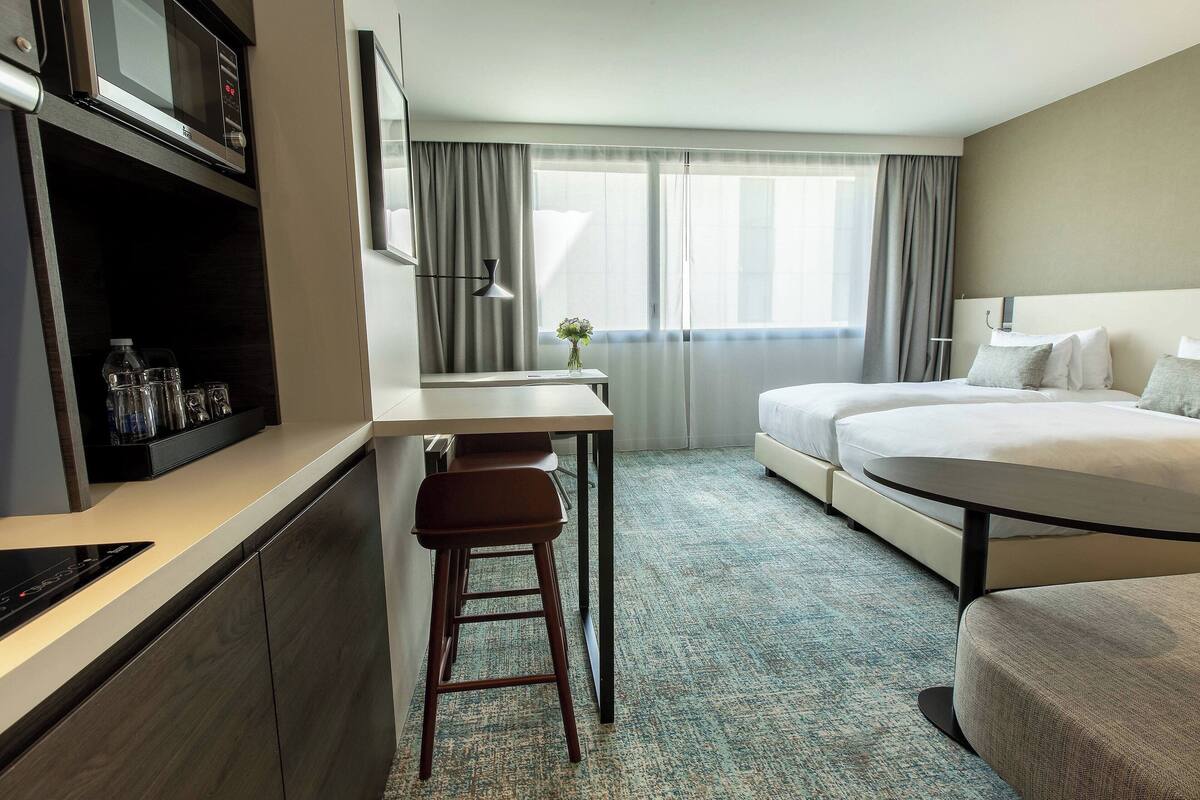 Une chambre d’hôtel comprenant un lit, un bureau avec une chaise, un four à micro-ondes, un distributeur d’eau et une fenêtre avec des rideaux.