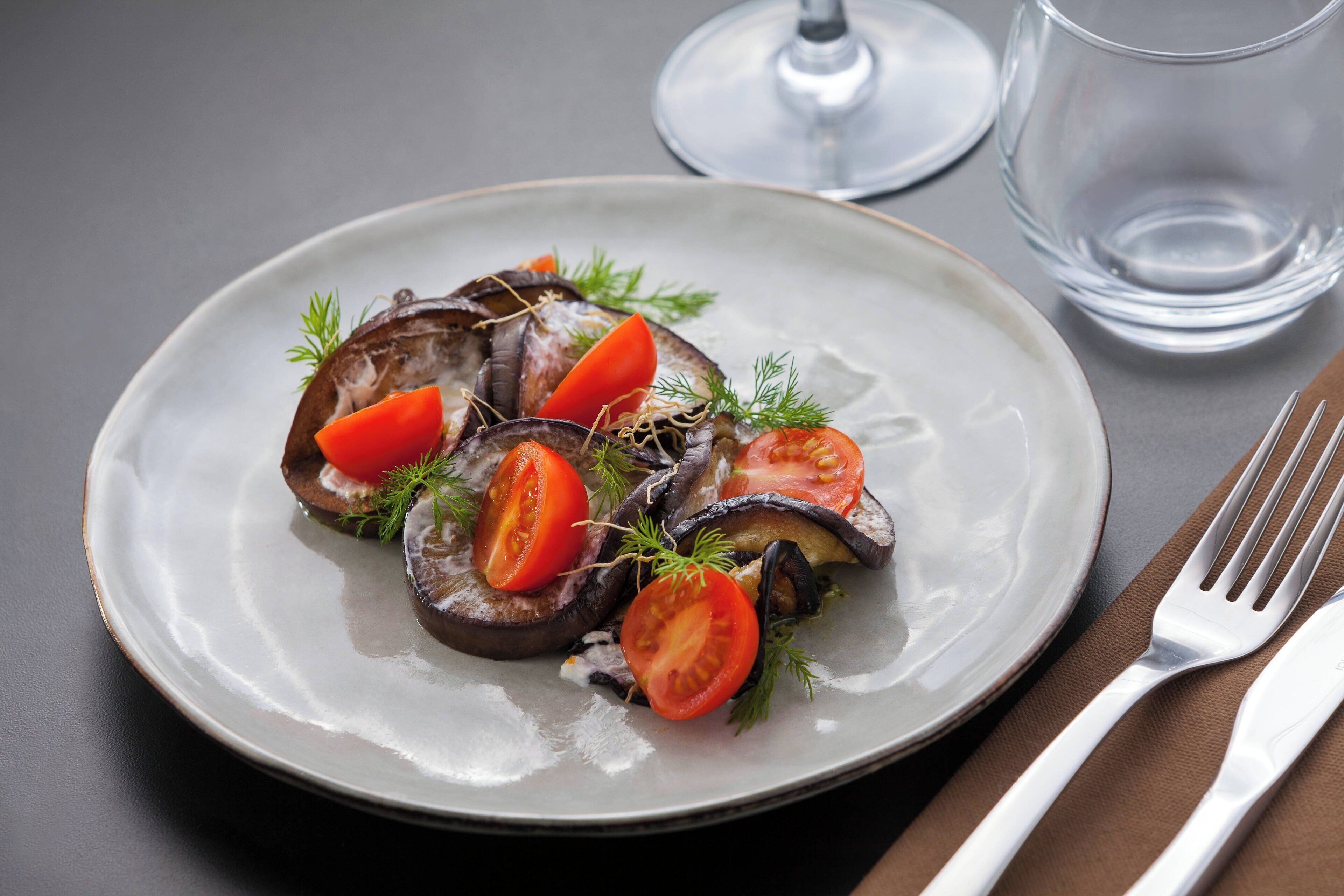 Un plato de berenjena en rodajas con tomates y hierbas sobre un plato blanco, acompañado de un tenedor y cuchillo sobre una servilleta marrón, y un vaso de agua sobre una mesa.