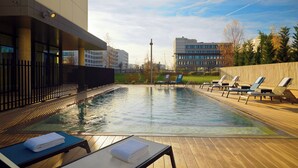 Une piscine entourée de bâtiments et d’arbres, avec des chaises longues et une terrasse en bois.
