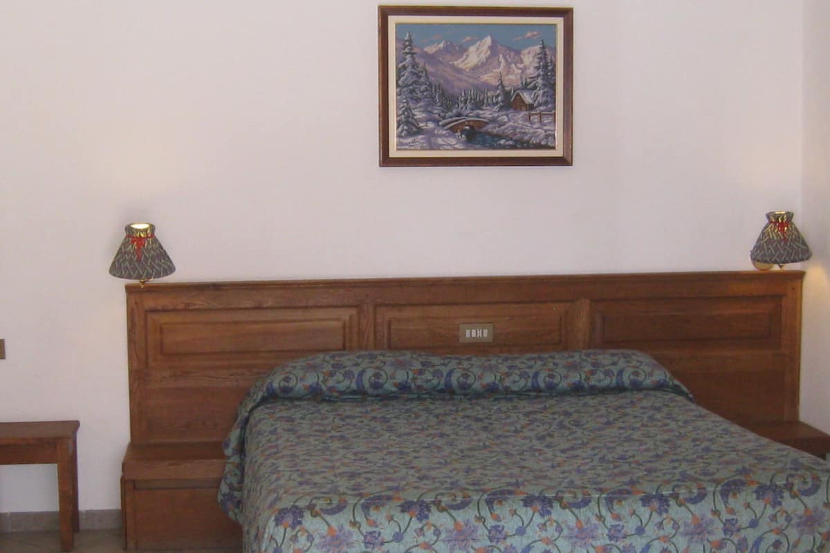 A bedroom with a wooden headboard, a bed with a patterned bedspread, two bedside lamps, and a framed picture of a snowy landscape on the wall.