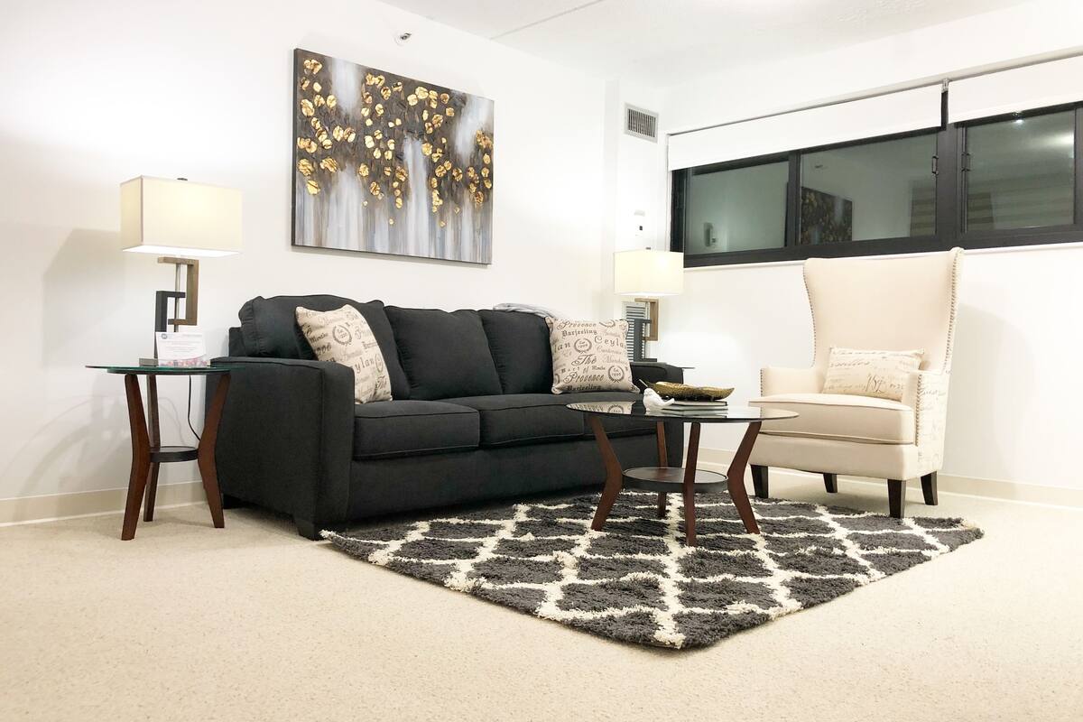 A living room with a black sofa, a white armchair, a glass coffee table, and a patterned rug.
