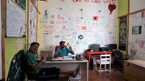 Una habitación con una pizarra llena de dibujos y escrituras de colores, un escritorio con dos personas trabajando y una lámpara de escritorio roja.