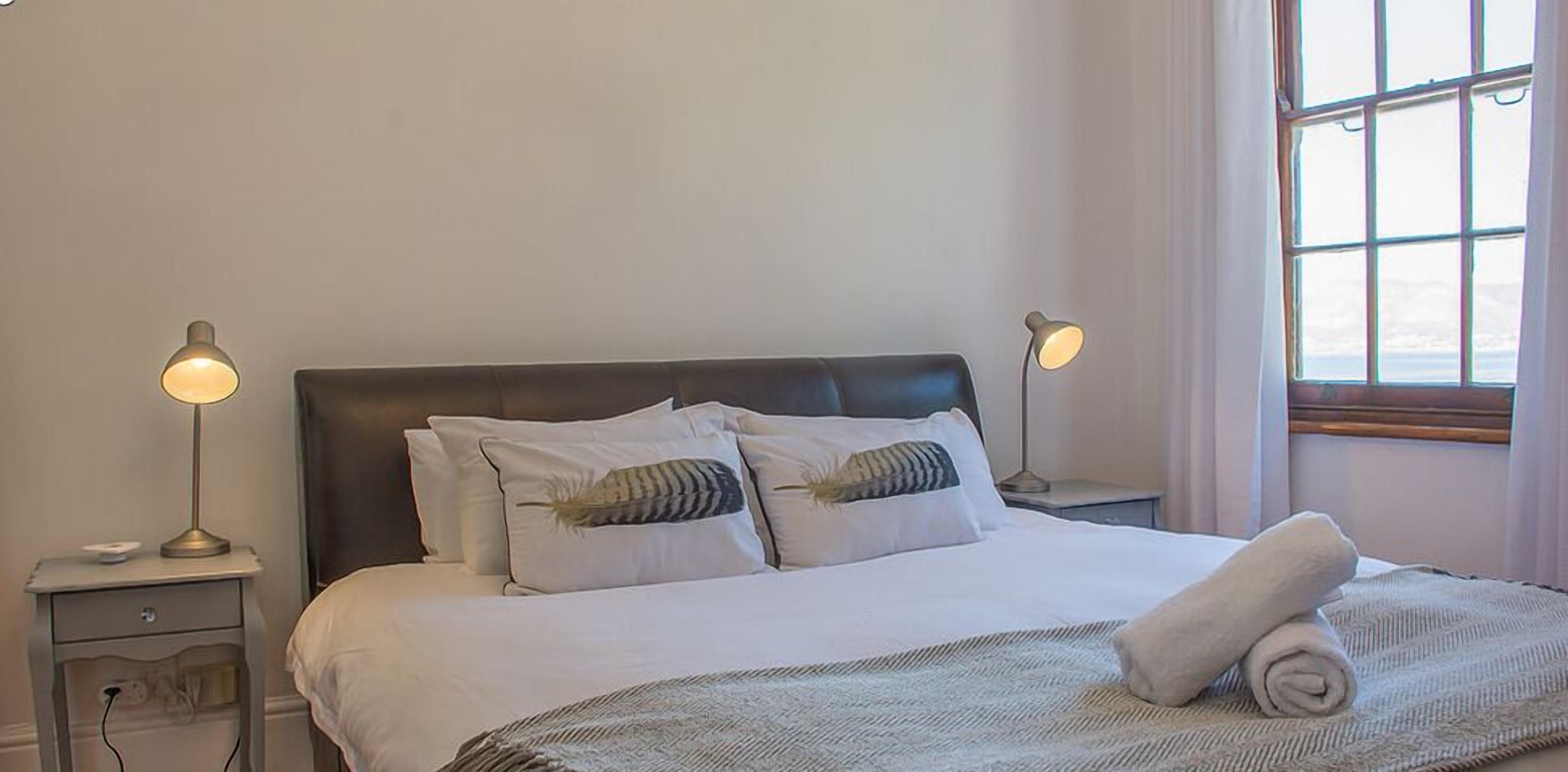 Cama bem arrumada com uma toalha enrolada, duas luminárias de cabeceira e uma janela com cortinas.