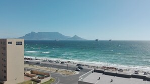 A coastal cityscape with a prominent building, a beach, and a mountainous backdrop. - The Waves, Living By the Beach (Cape Town)