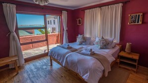 A bedroom with a large bed, two bedside tables, a wooden bench, and a view of a red building and greenery through a large window. - Island Cottage Guesthouse (Luderitz)