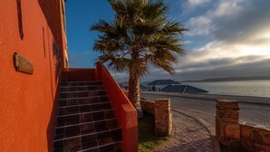 A staircase leading to a building with a red exterior, a palm tree, and a view of the sea and distant islands. - Island Cottage Guesthouse (Luderitz)