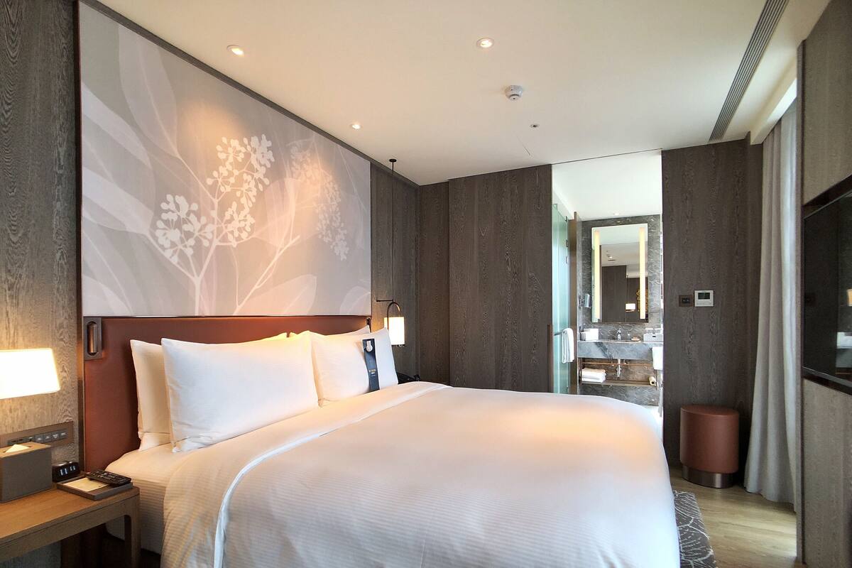 A hotel room with a large bed, a nightstand, a wall-mounted lamp, and a bathroom visible through an open door.
