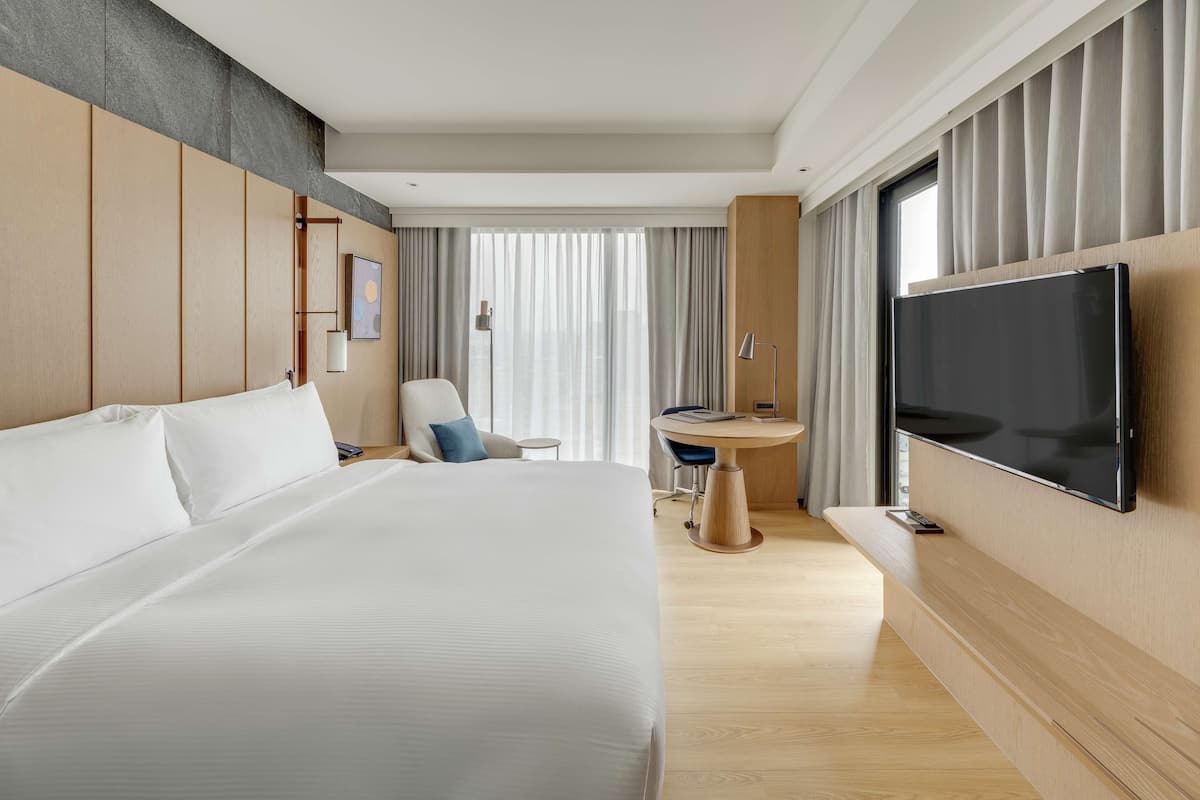 A modern hotel room with a large bed, a desk, a TV, and a view of the city through large windows.