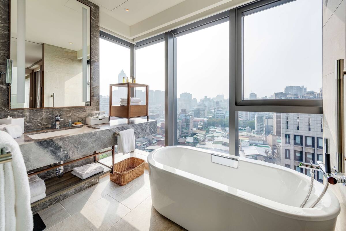 A modern bathroom with a large bathtub, a marble vanity, a large mirror, and a view of the city through floor-to-ceiling windows.