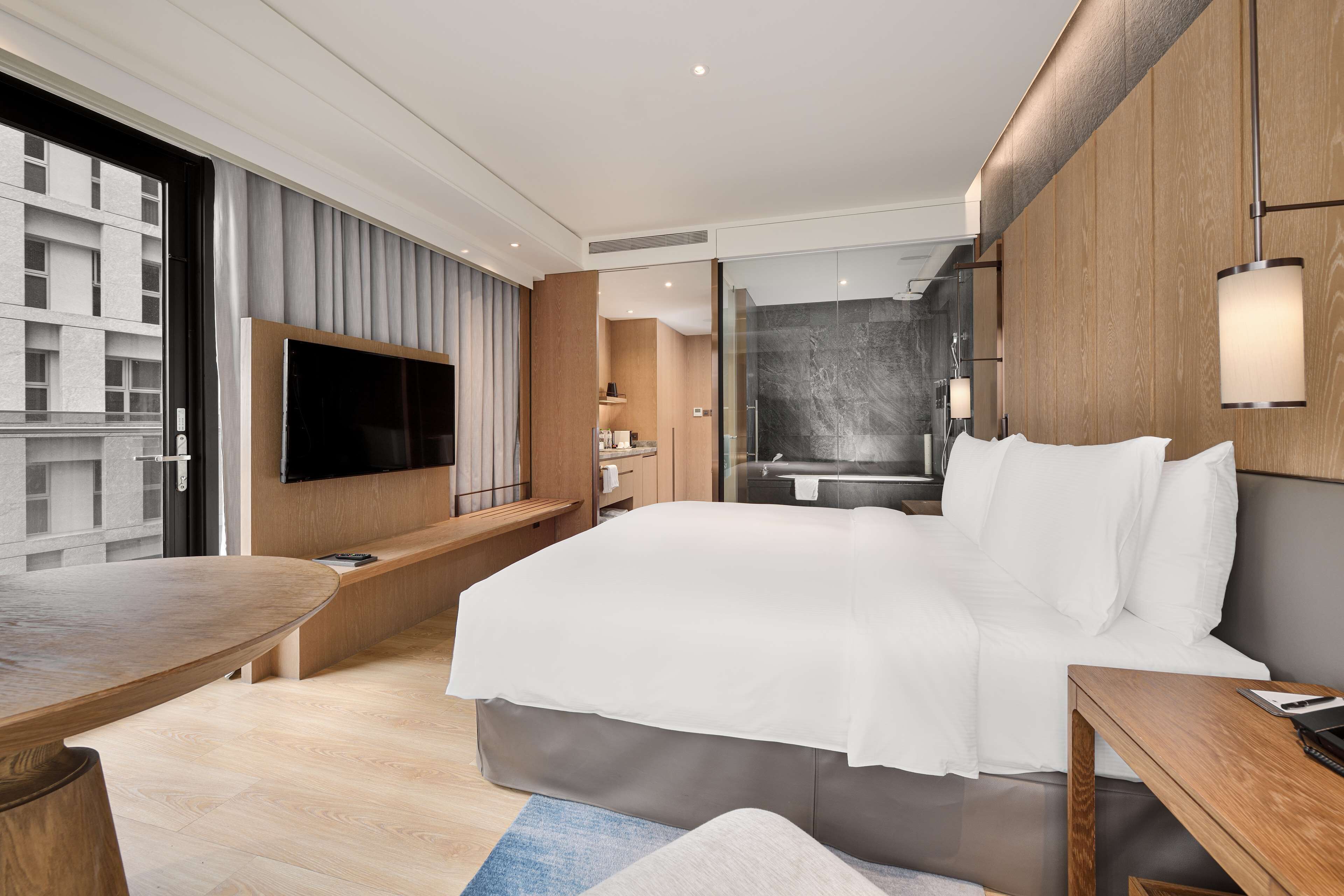 A modern hotel room with a large bed, a TV mounted on the wall, a wooden desk, and a view of the city through the window.