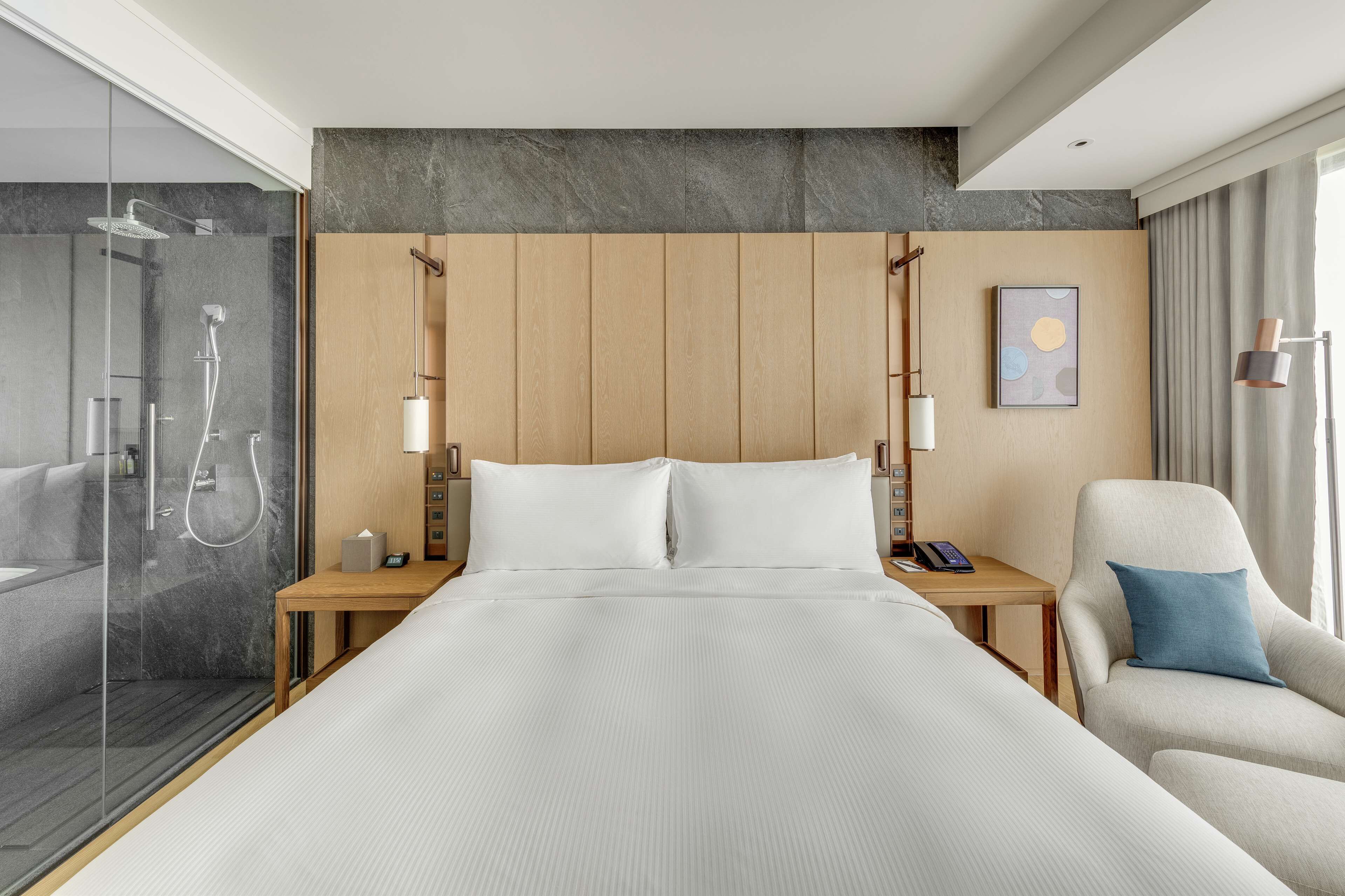 Quarto de hotel com cama grande, mesas de cabeceira, uma cadeira, área de chuveiro e um quadro na parede.