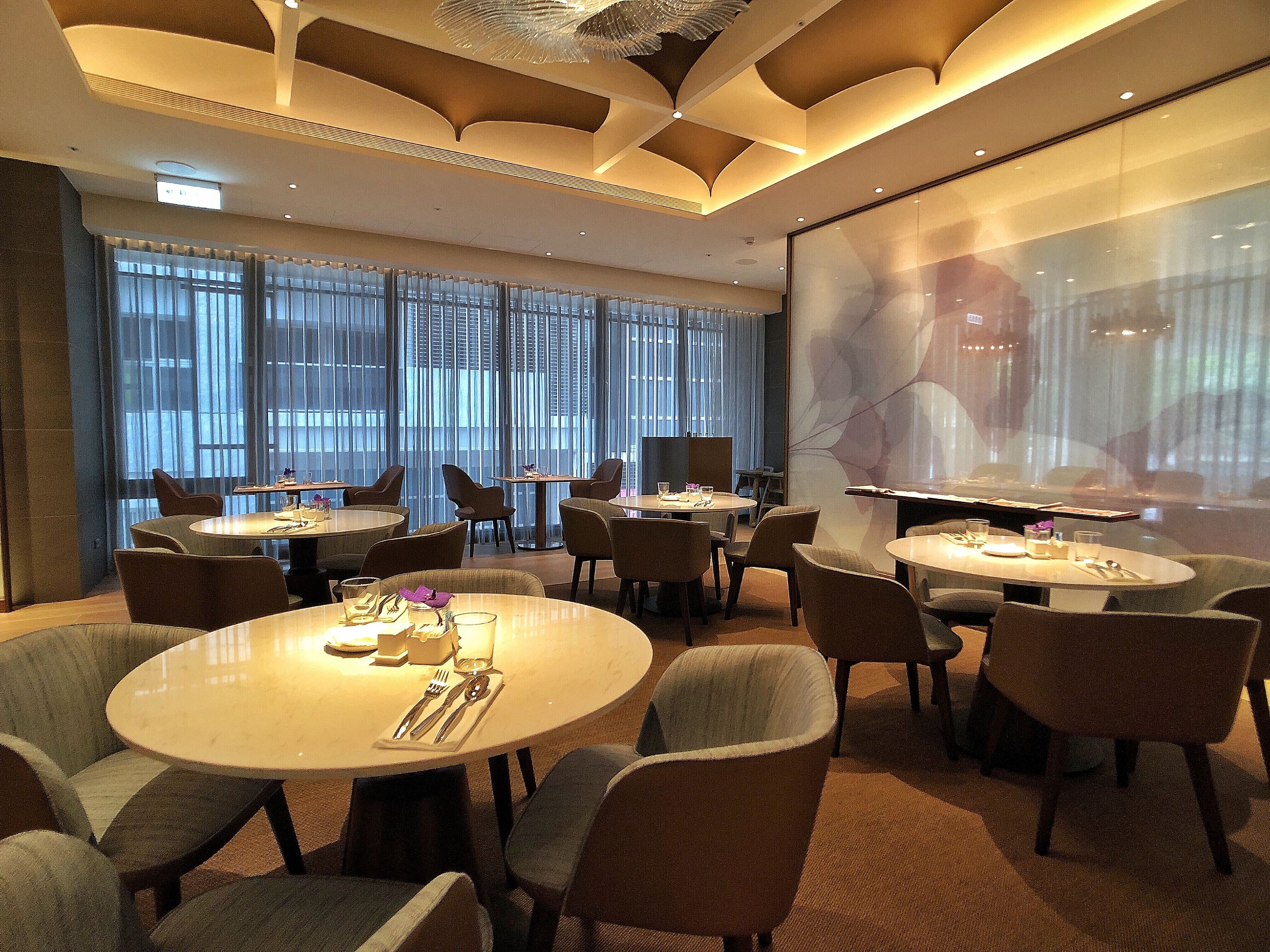 A modern hotel room with round tables and chairs, large windows with curtains, and a projection screen on the wall.
