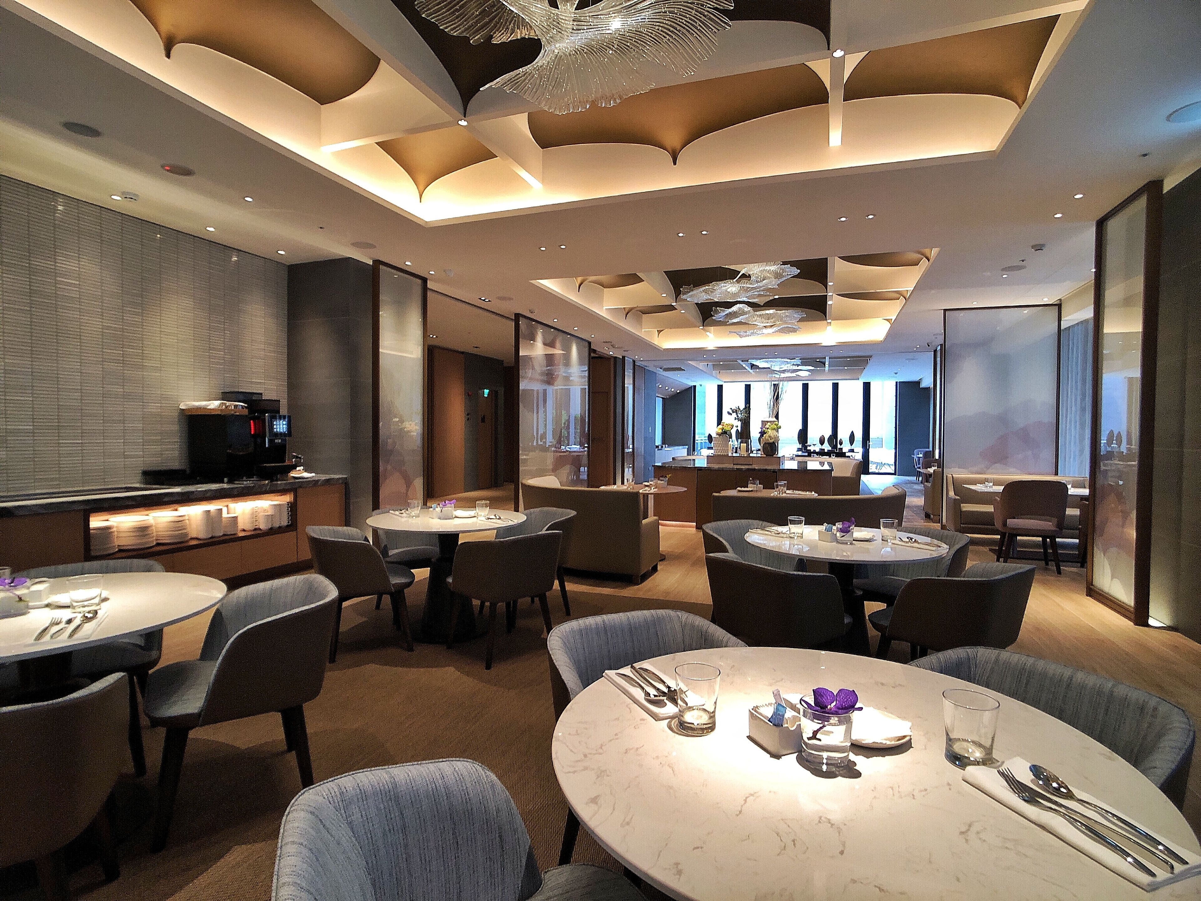 A modern hotel lobby with round tables, comfortable seating, a coffee machine, and a view of the city through large windows.