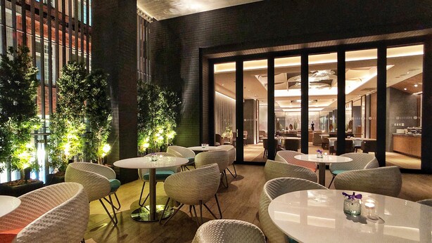 A modern indoor dining area with round tables and wicker chairs, featuring a brick wall and indoor plants. - DoubleTree by Hilton Taipei Zhongshan (Taipei)