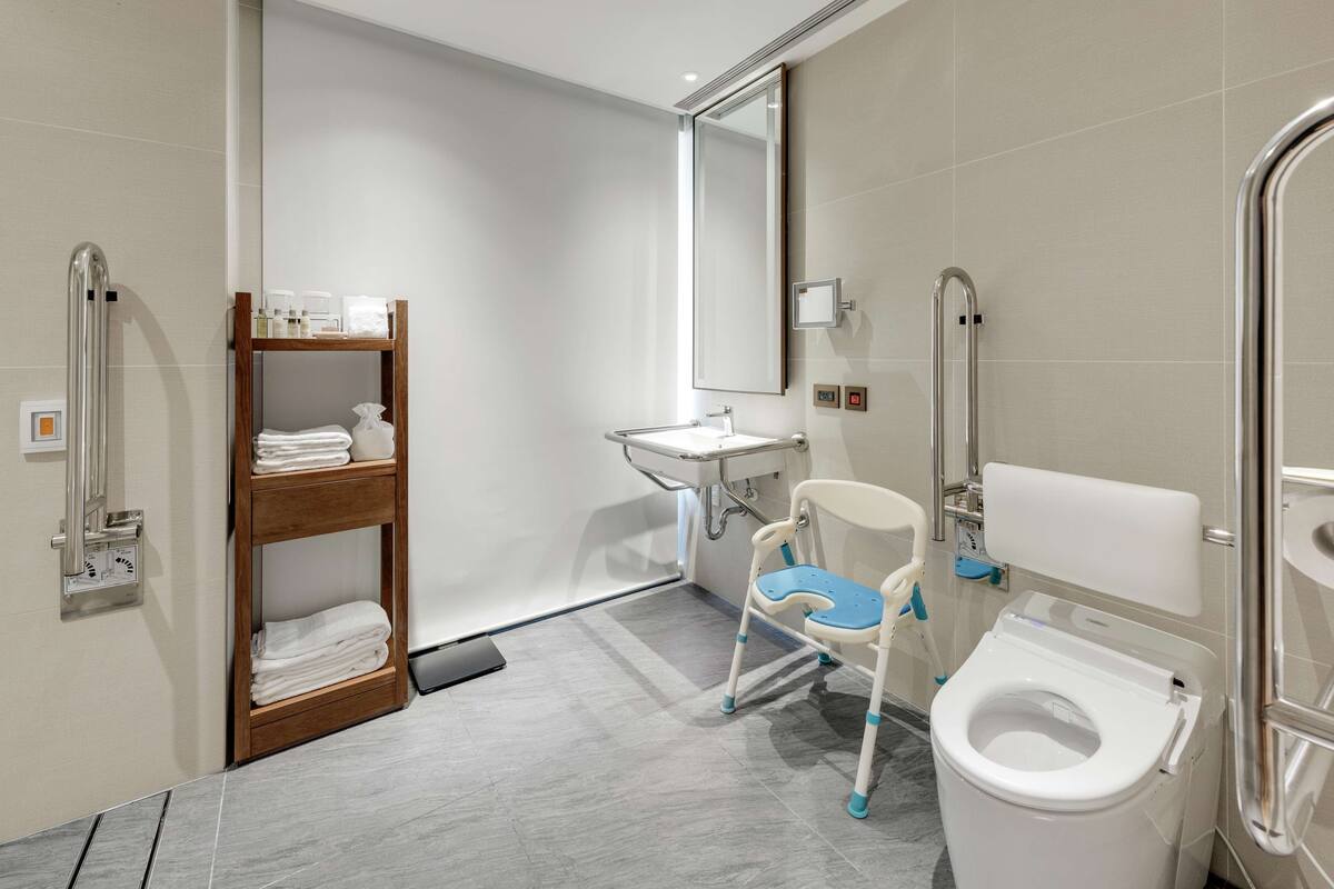 A modern bathroom with a white sink, a toilet with a safety seat, a wooden shelf with towels, and a handrail.