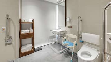 A modern bathroom with a white sink, a toilet with a safety seat, a wooden shelf with towels, and a handrail.