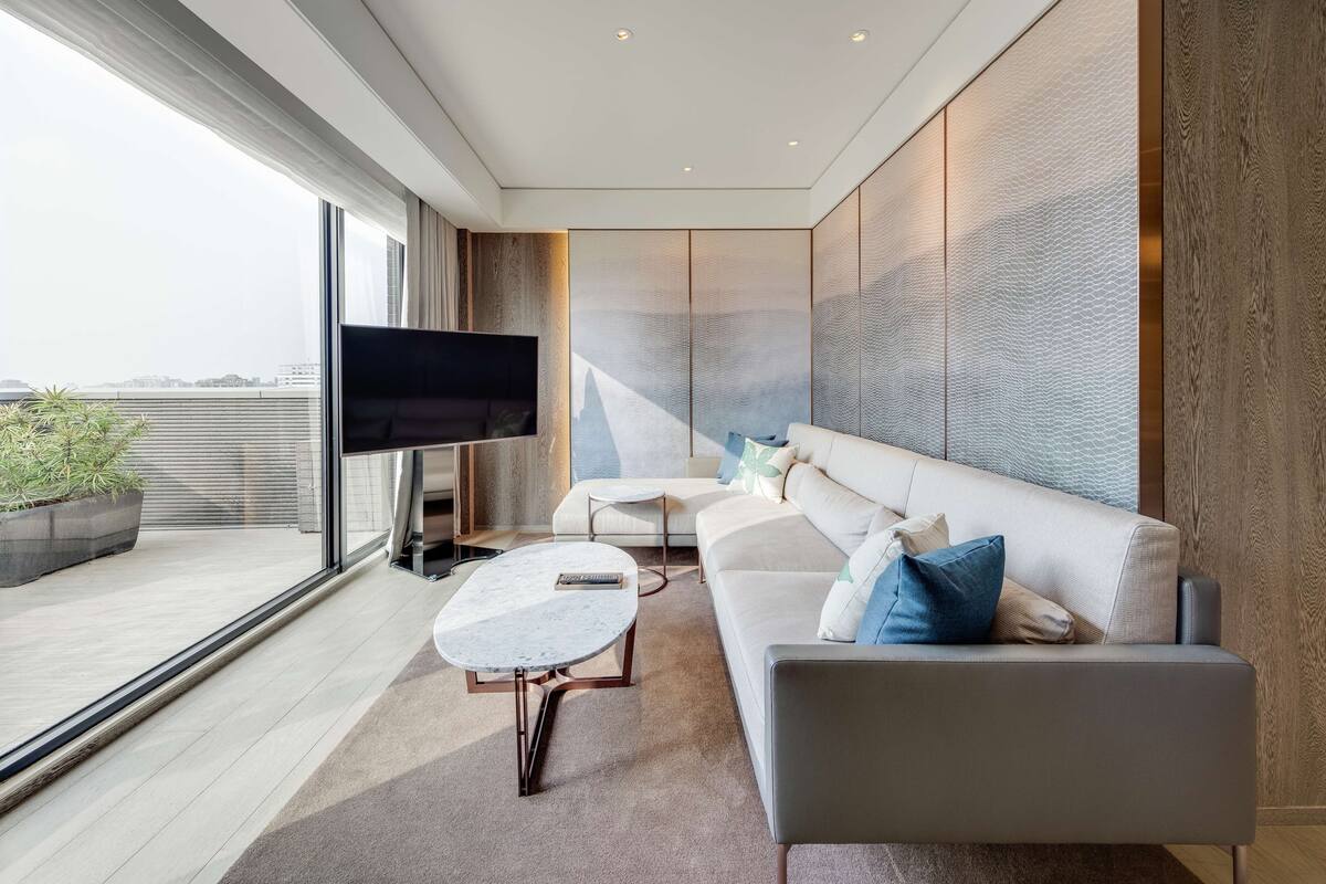A modern living room with a large flat-screen TV, a sofa, a coffee table, and a view of the city through large windows.