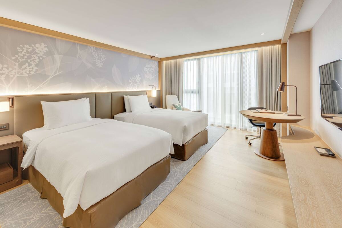 A hotel room with two beds, a desk, a chair, a TV, and a large window with curtains.