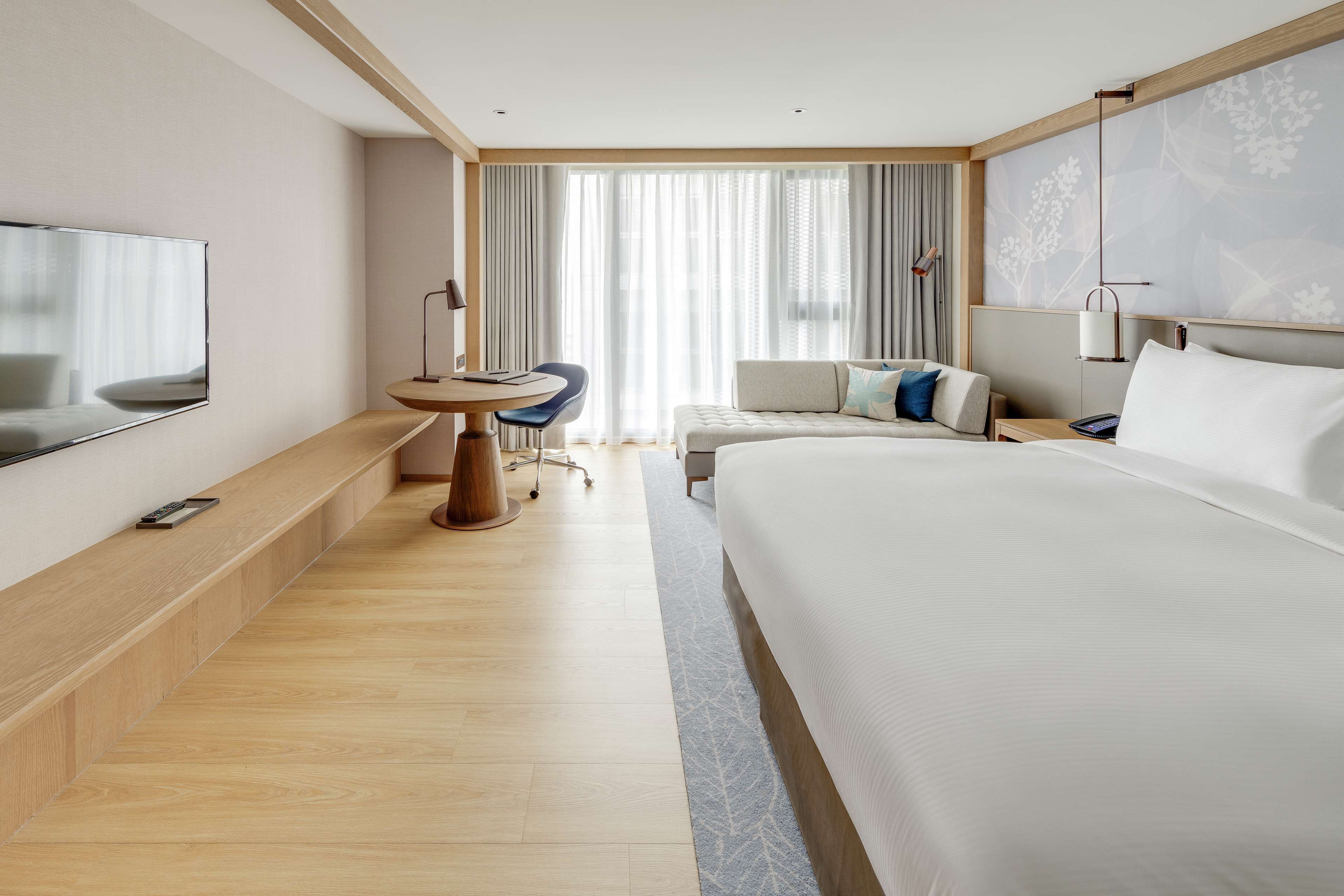 A modern hotel room with a large bed, a desk with a chair, a TV mounted on the wall, and a large window with curtains.