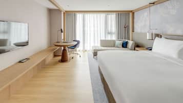 A modern hotel room with a large bed, a desk with a chair, a TV mounted on the wall, and a large window with curtains.