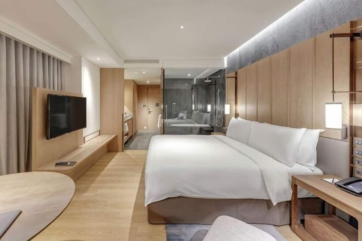 A modern hotel room with a large bed, a TV, a desk, and a bathroom visible in the background.