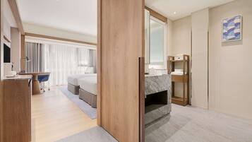 A modern hotel room with a wooden floor, a large bed, a desk with a chair, a vanity with a mirror, and a large window with curtains.