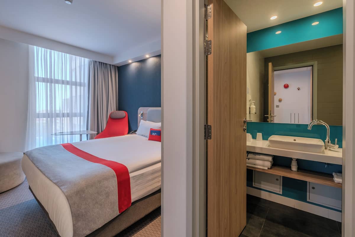 A hotel room with a bed, a red chair, a bathroom with a sink, and a mirror.