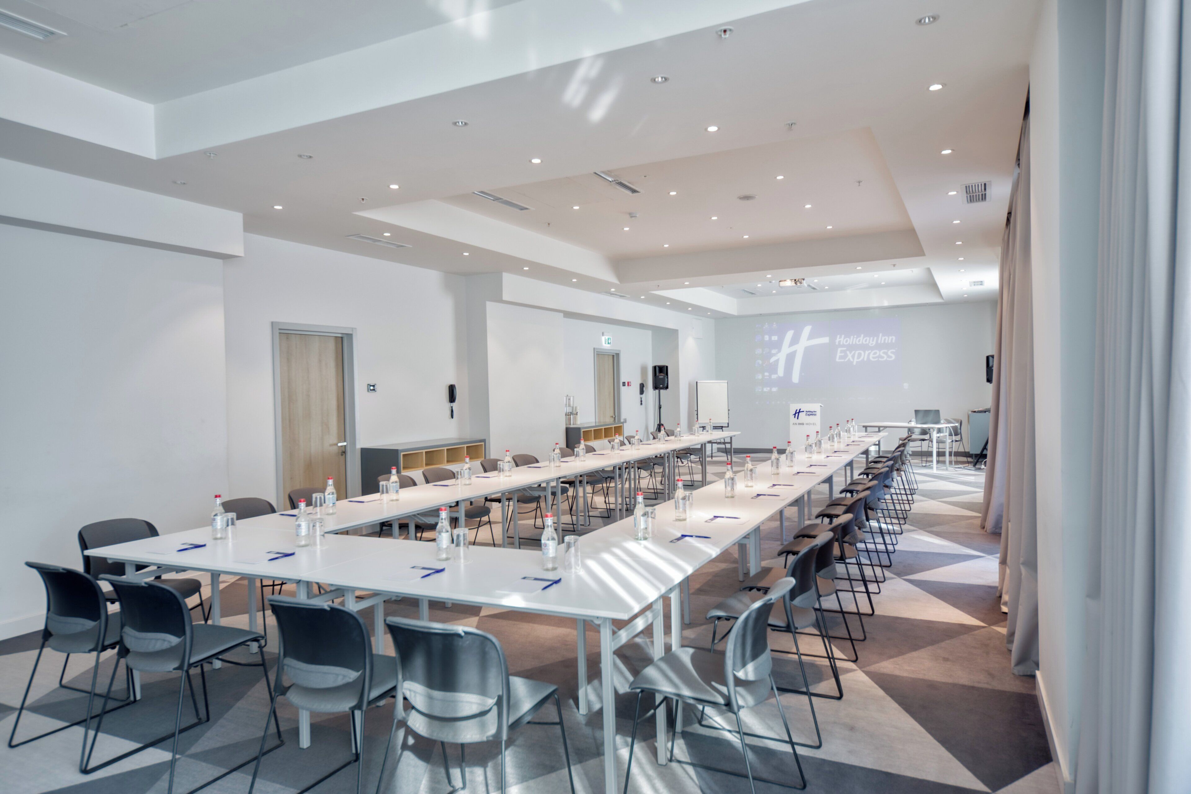 Una sala de conferencias con una larga mesa, sillas y una pantalla de proyección mostrando 'Holiday Inn Express'.